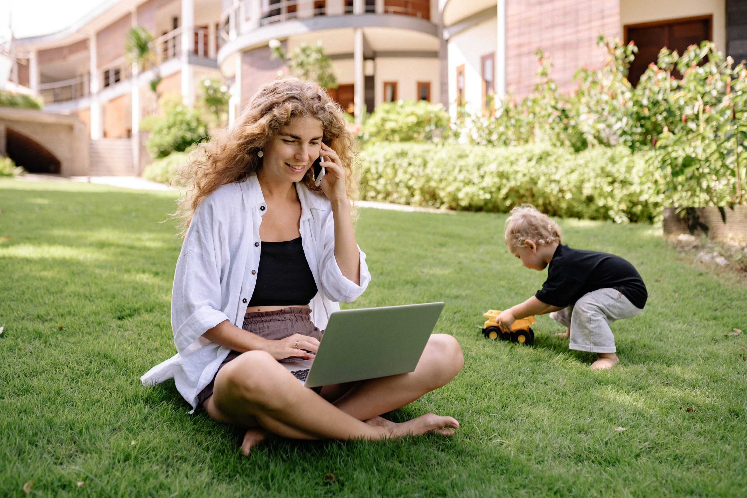 a woman on the phone on her computer while her son plays on the grass next to her
