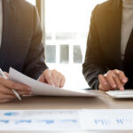 Two professionals reviewing a document and calculating figures at a desk with financial charts