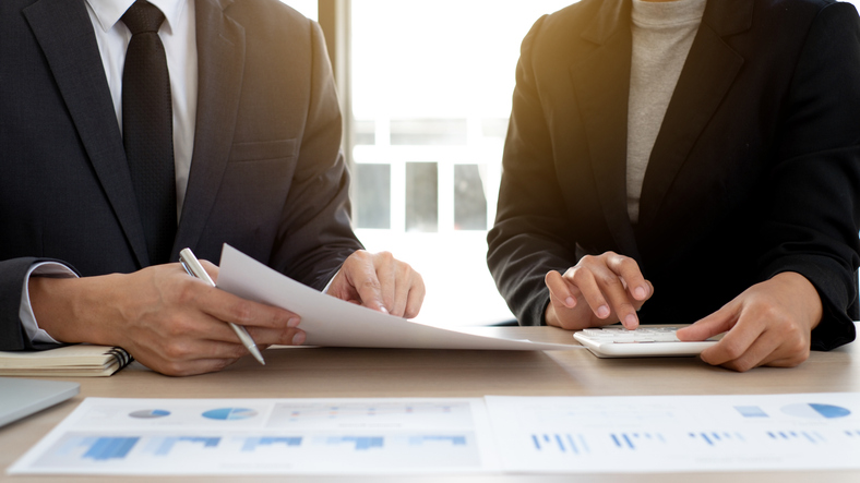 Two professionals reviewing a document and calculating figures at a desk with financial charts
